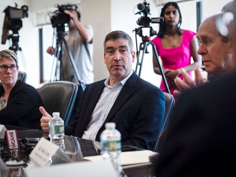 A man in a suit speaks during a meeting while seated at a conference table. Two people, one in a pink dress and another in a casual outfit, operate cameras in the background.
