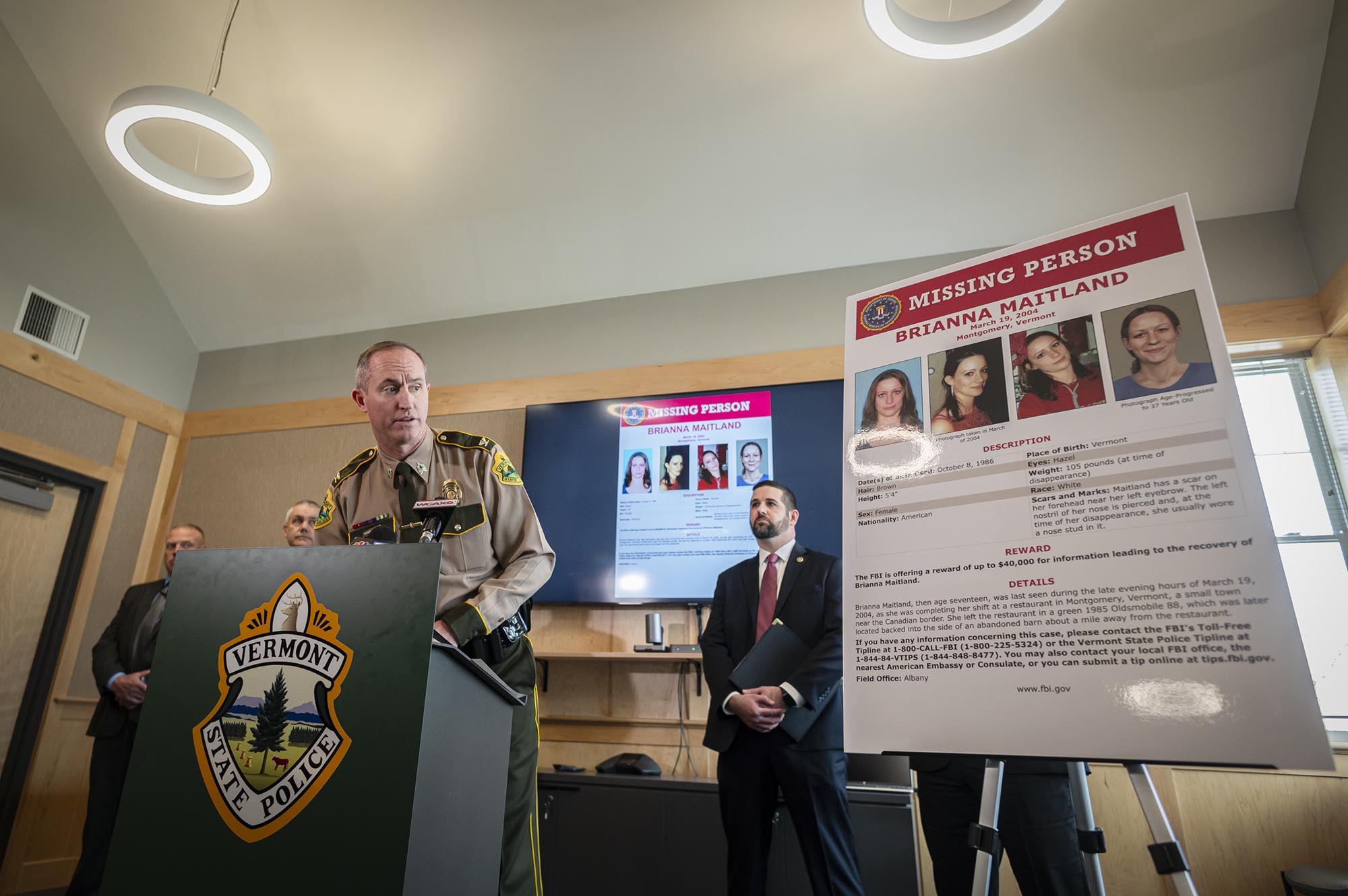 A law enforcement officer speaking at a podium during a press conference with a missing person poster in the background.