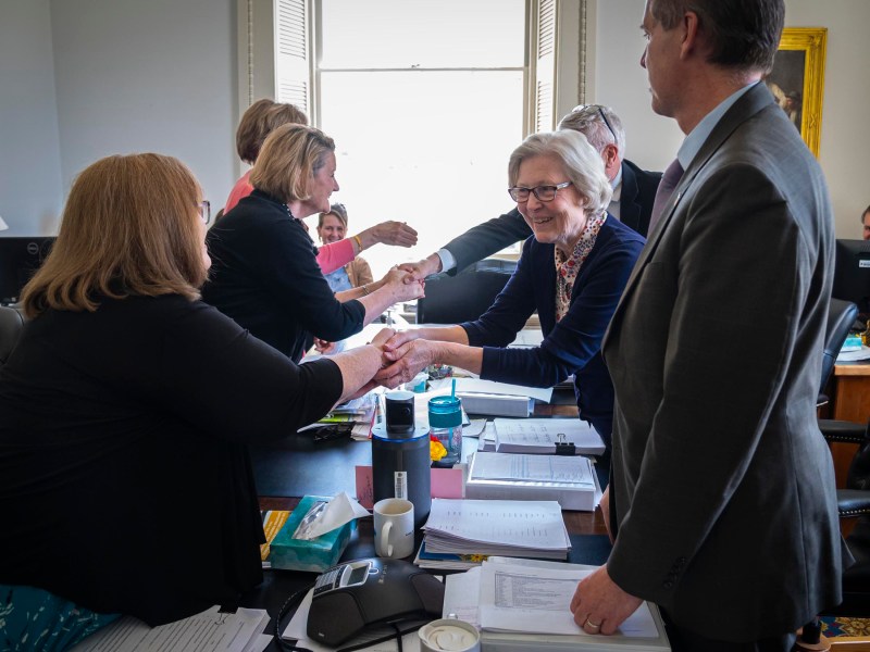 Business professionals shaking hands across a table in a busy office, with colleagues smiling and watching.