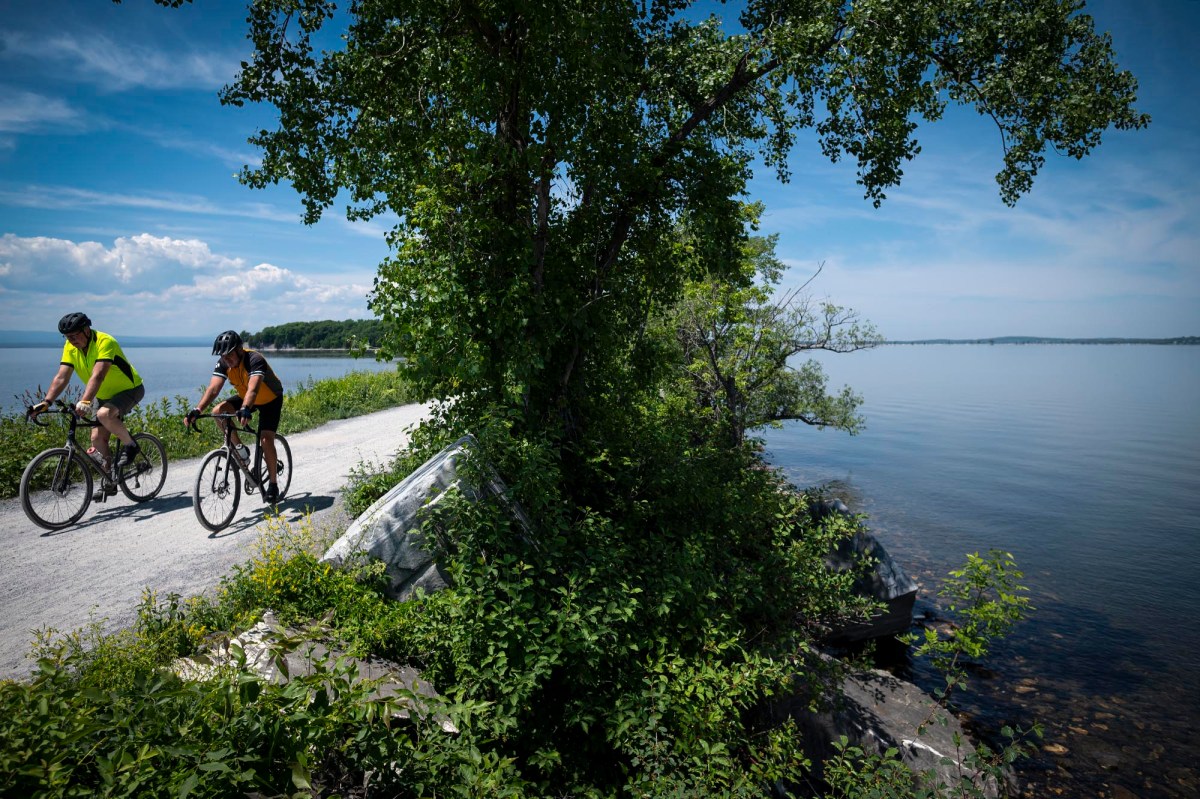 Two cyclists ride on a gravel path by a large tree and a body of water. The sky is clear with a few scattered clouds.