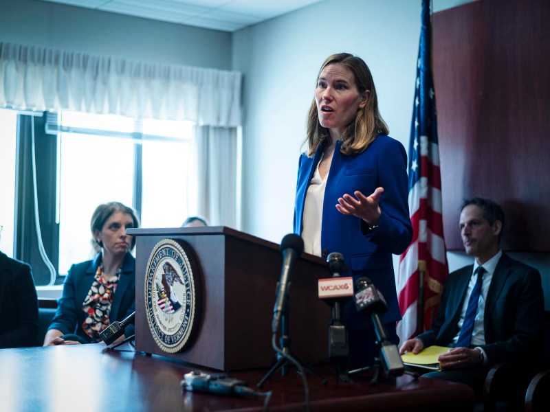 A woman speaks at a podium in a conference room with microphones and an american flag, while several people listen.