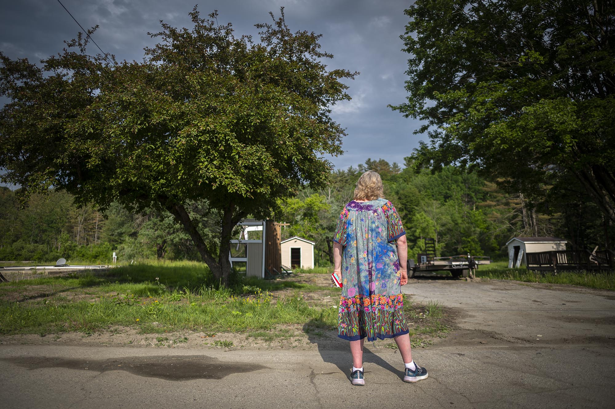 An older woman wearing a colorful dress stands on a paved area, looking at a small shed surrounded by greenery and trees on a sunny day.
