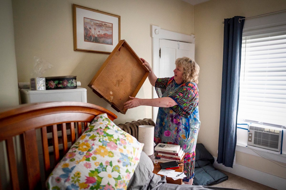An elderly woman in a colorful dress inspects an empty dresser drawer in a bedroom. The room has a bed with floral pillows, a painting on the wall, and a window with closed blinds.