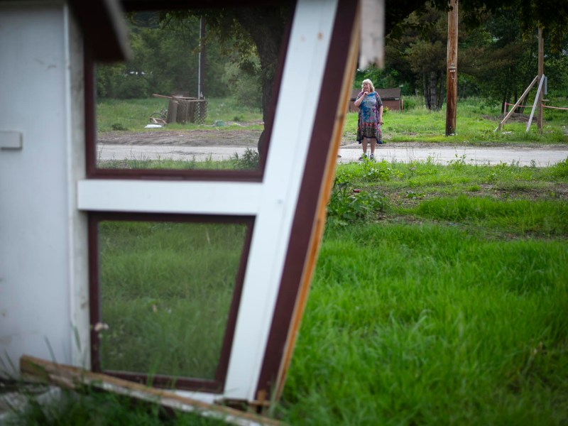 A person stands on a grassy roadside, seen through the frame of a tilted, broken structure in the foreground.