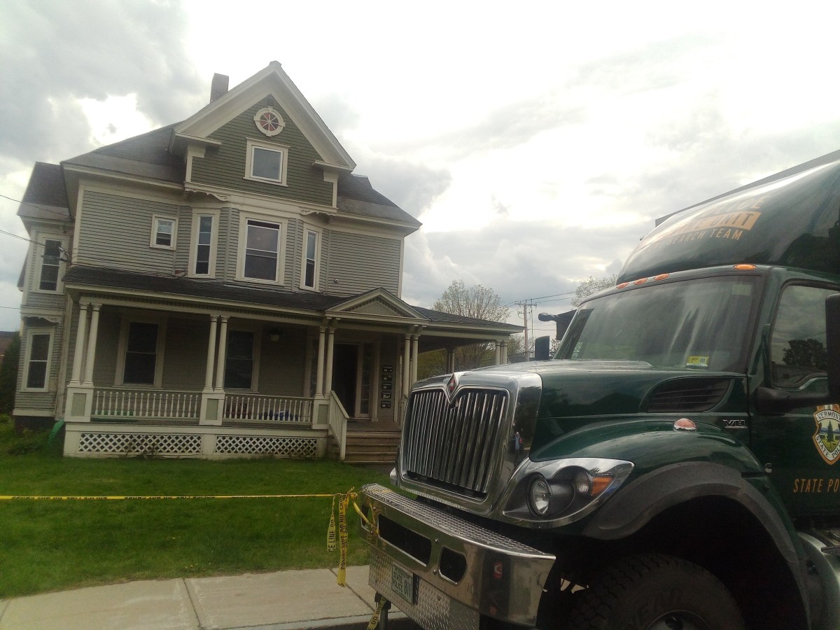 A green state police vehicle is parked in front of a large two-story house with a wraparound porch, cordoned off with yellow police tape.