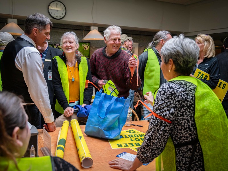 Senior man laughing joyfully during a conversation with a group at a community event table, surrounded by people wearing yellow vests and "tap es" badges.