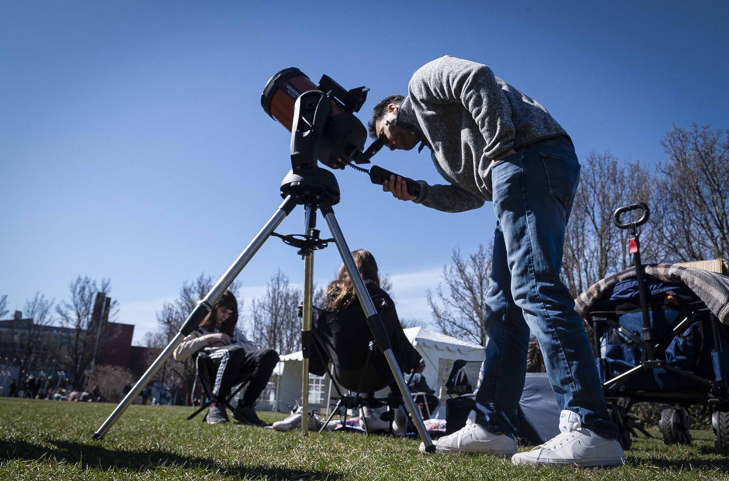 A person looking through a telescope during the daytime with others sitting on the grass nearby.