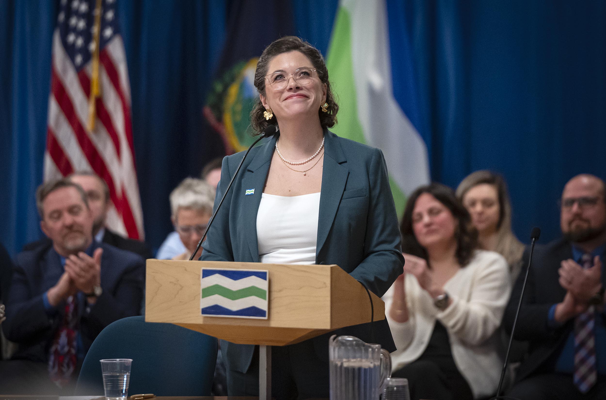 A woman standing at a podium with a smiling expression while people seated in the background are applauding.