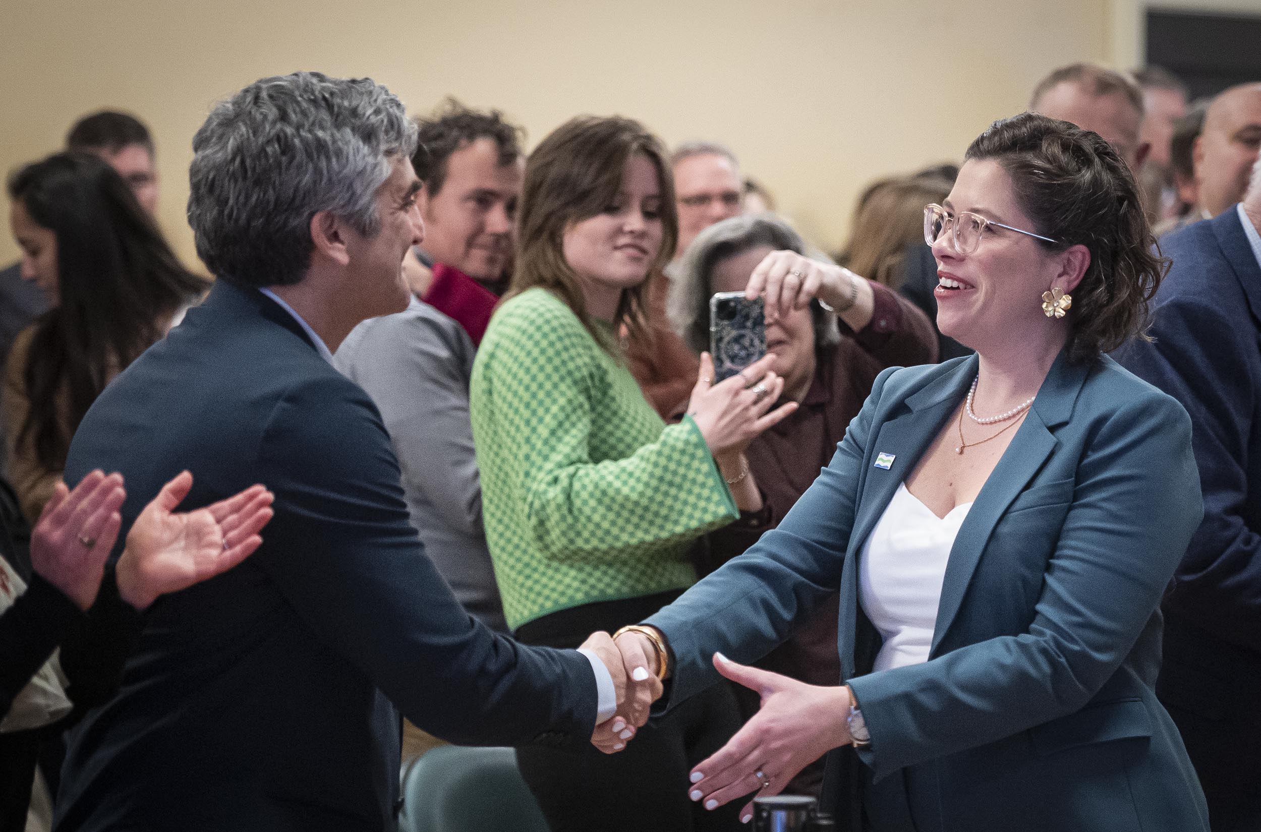 Two individuals shaking hands and smiling at a formal event while others in the background observe and take photographs.
