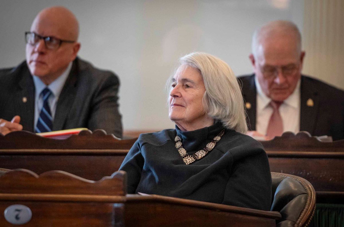 A woman with gray hair, wearing a black outfit and a decorative brooch, sitting attentively at a wooden desk in a formal meeting room, with two men seated behind her.