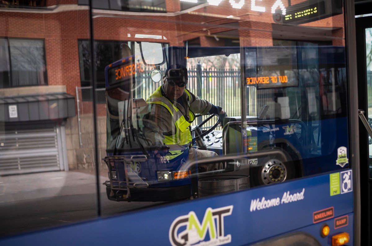 Bus driver operating a vehicle viewed through the windshield with reflections of the surrounding area visible.