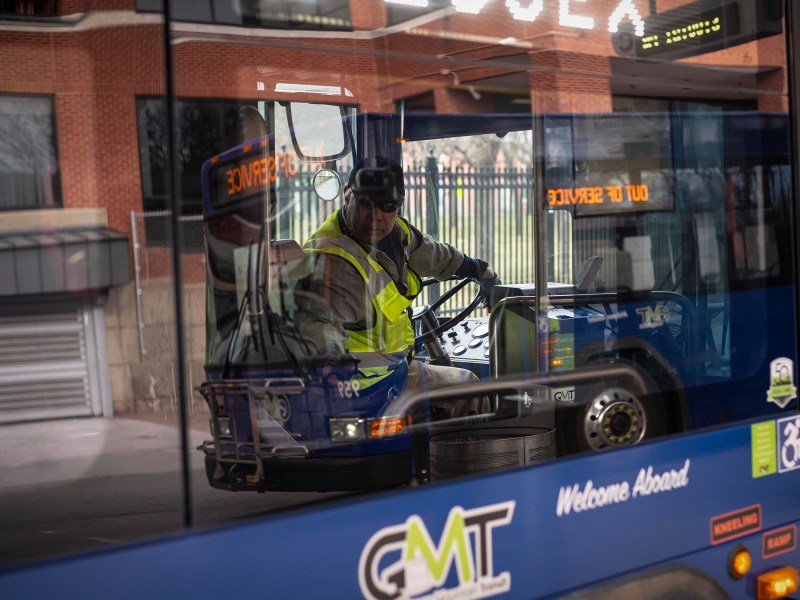 Bus driver operating a vehicle viewed through the windshield with reflections of the surrounding area visible.