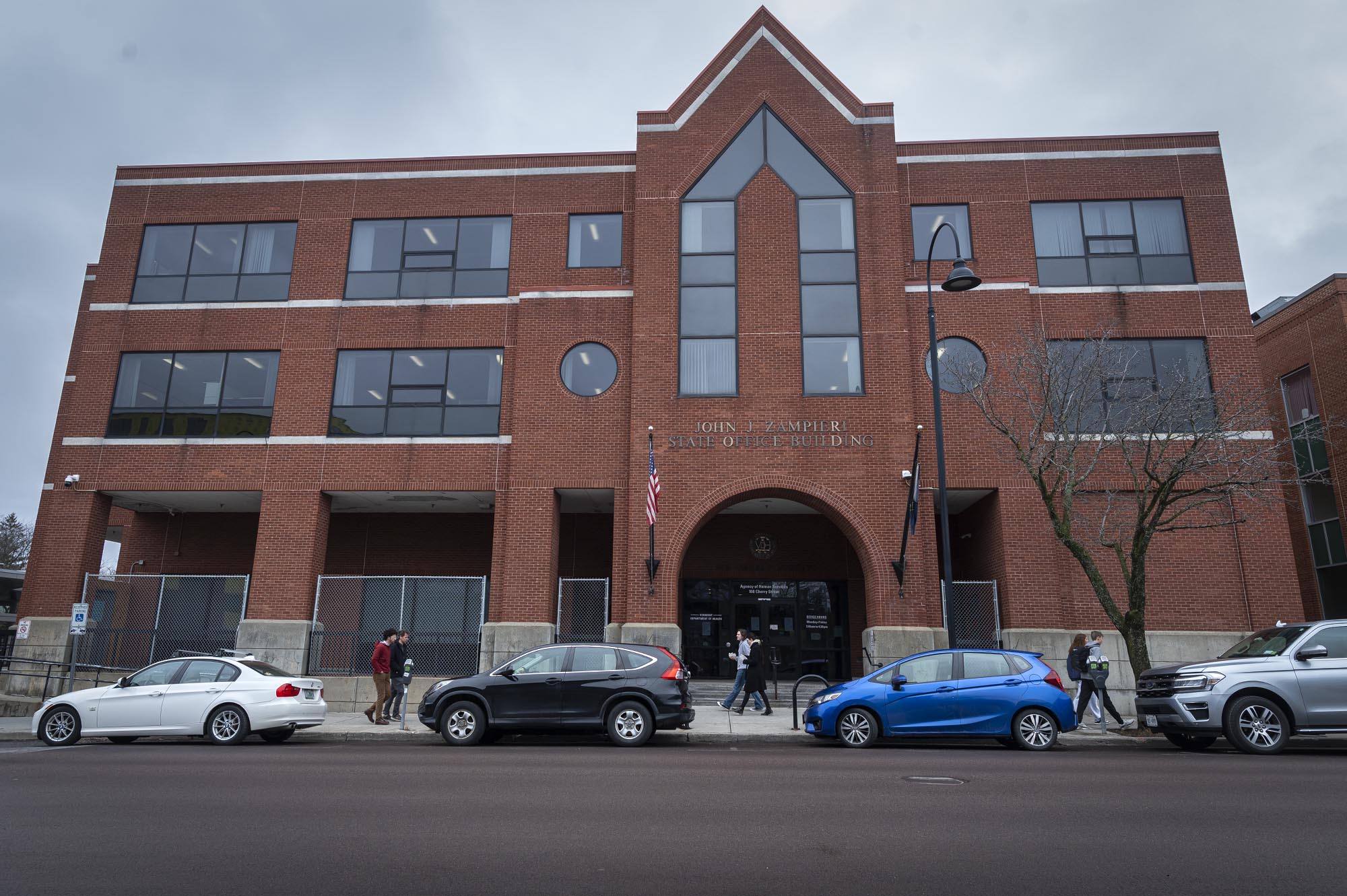 Modern brick building with people walking by and parked cars in the foreground.