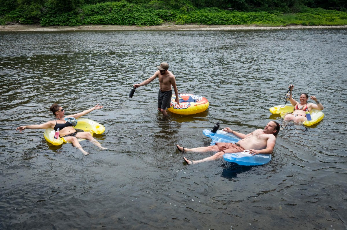 Four people relax on inflatable floats in a river, holding bottles. One person stands in the shallow water with a bottle and a hat. The riverbank is lush with greenery in the background.