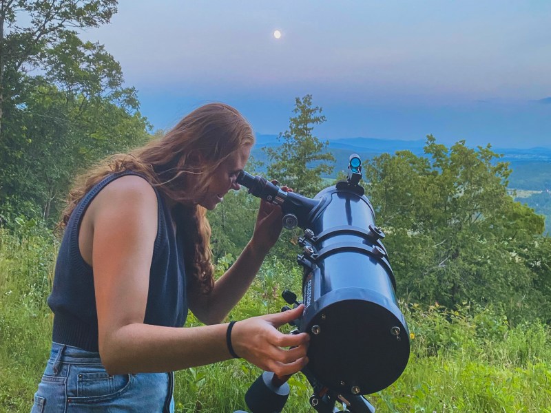 A woman looking through a telescope on a hillside.