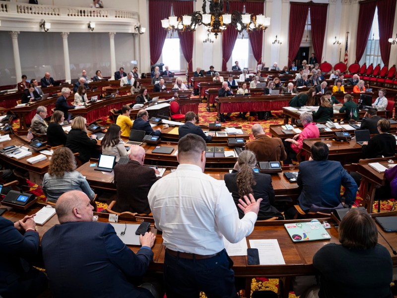 A busy legislative assembly session in a grand chamber, with members engaged in discussion and laptop use.