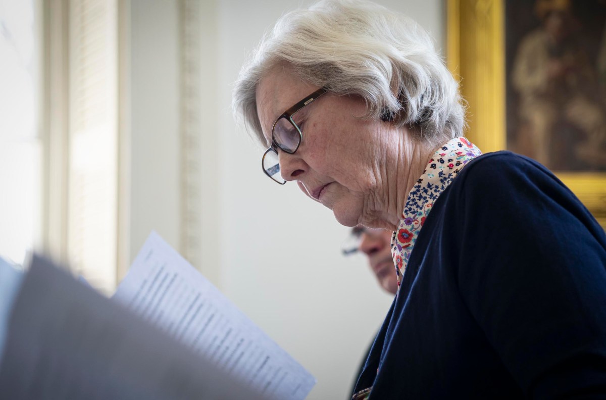 An elderly woman with glasses, wearing a scarf, intently reads a document in a room with soft lighting.
