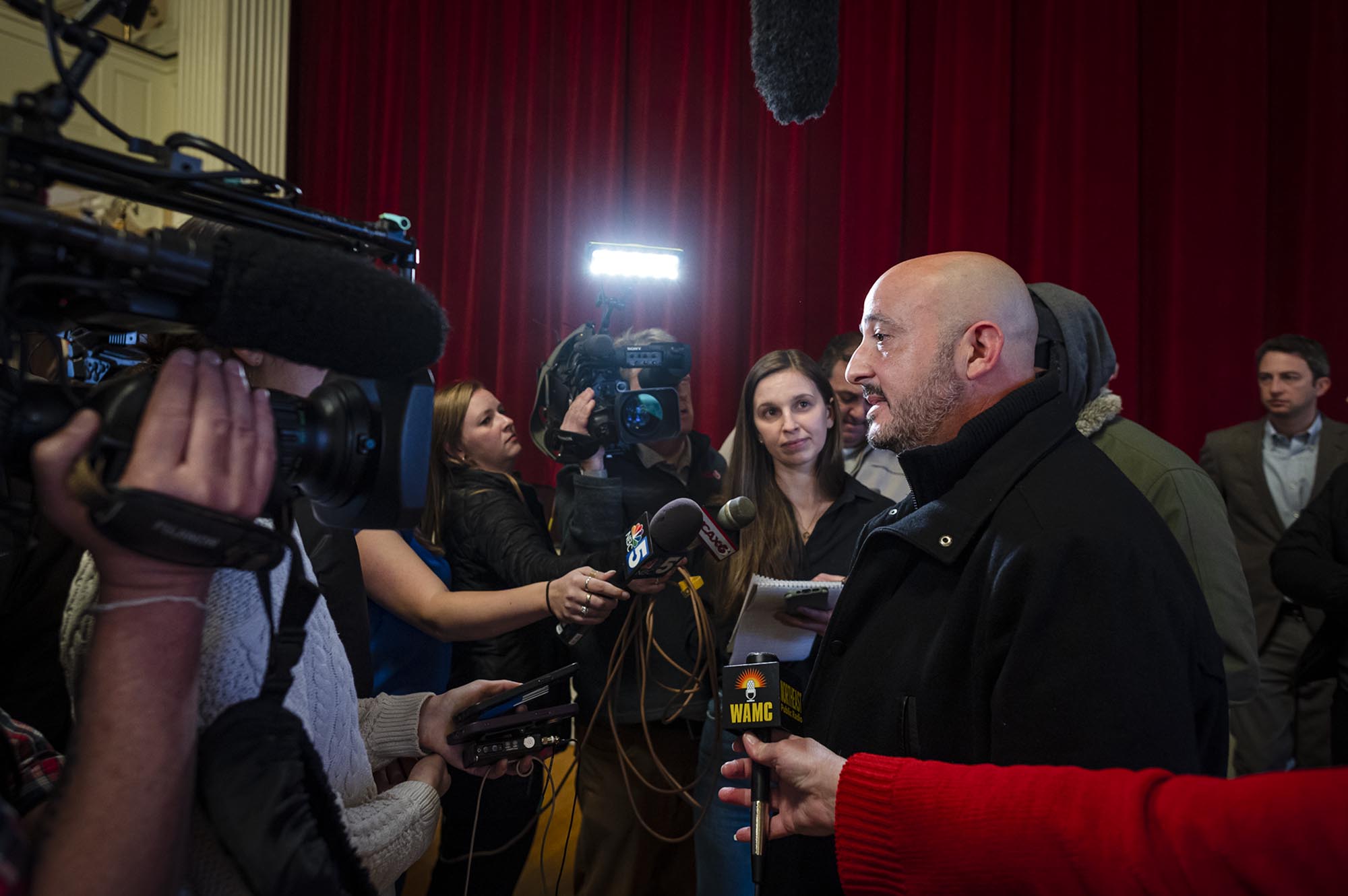 A bald man talking to reporters in front of a crowd.