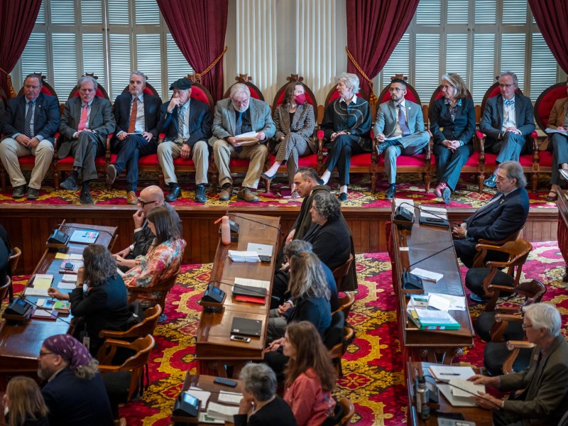 A group of people seated in a formal meeting room, some appearing attentive while others are distracted or tired.