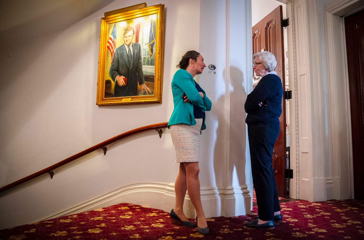 Two women engaging in conversation in a hallway with a portrait hanging on the wall.