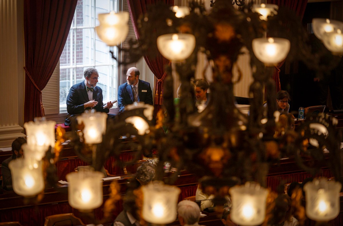 Two men conversing in a grand, dimly lit hall with large windows and ornate metal candelabras emitting soft light foreground.