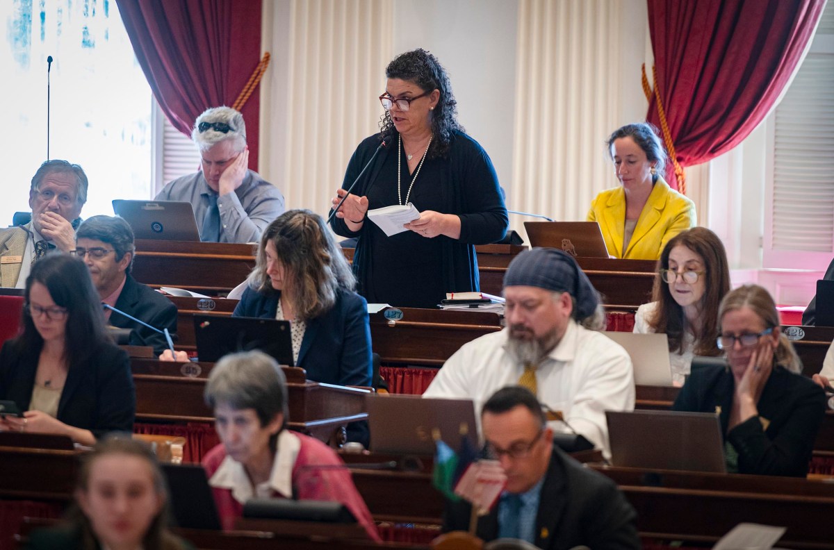 A woman stands speaking and reading from a note card in a legislative chamber filled with seated colleagues.