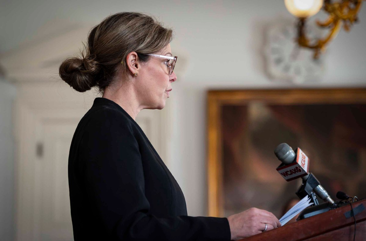 A woman in glasses and a black outfit speaks at a podium with microphones, in a room with elegant decor.