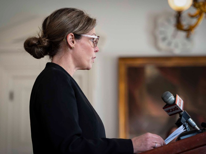 A woman in glasses and a black outfit speaks at a podium with microphones, in a room with elegant decor.
