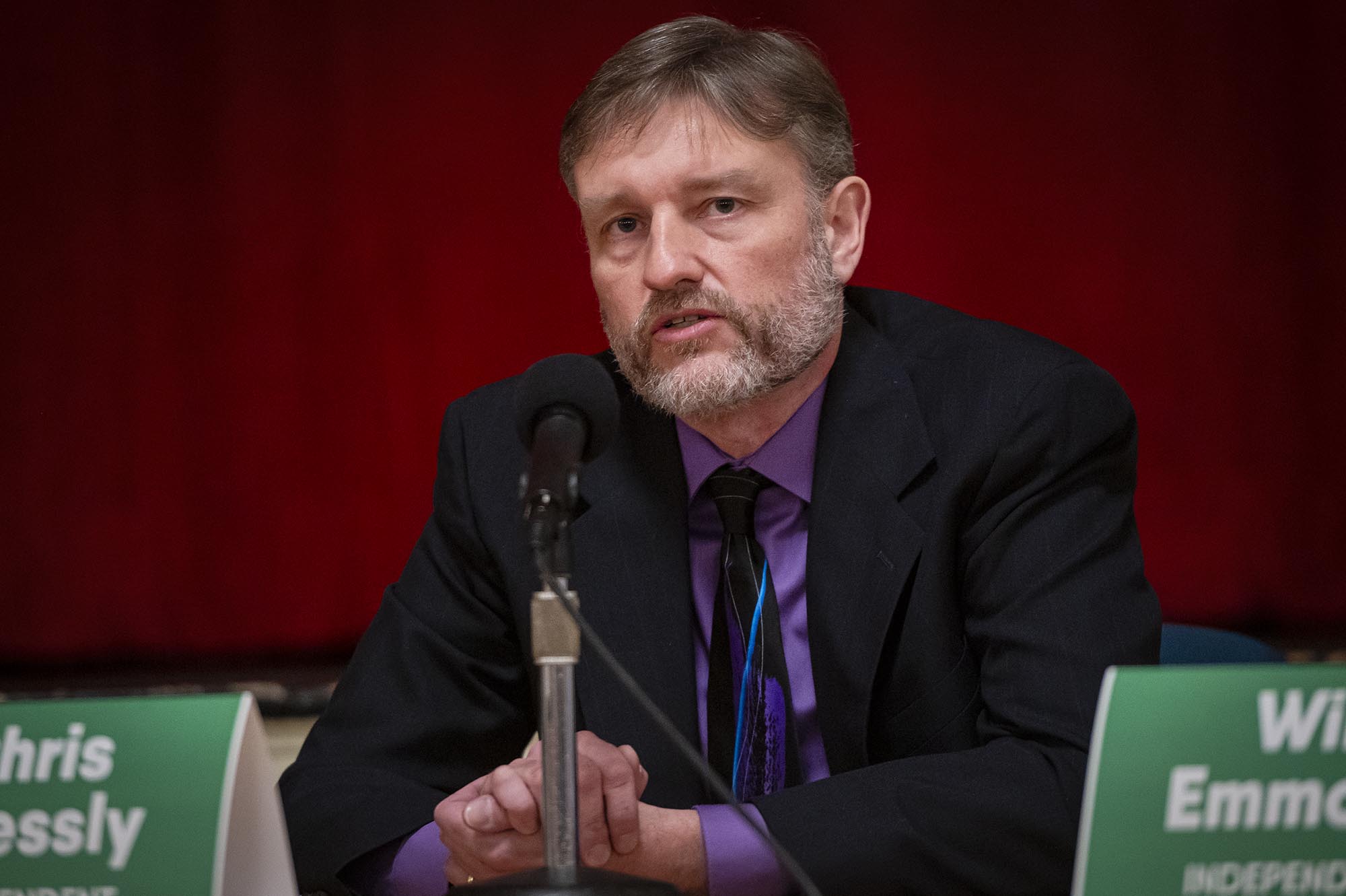 A man in a suit sits at a microphone.