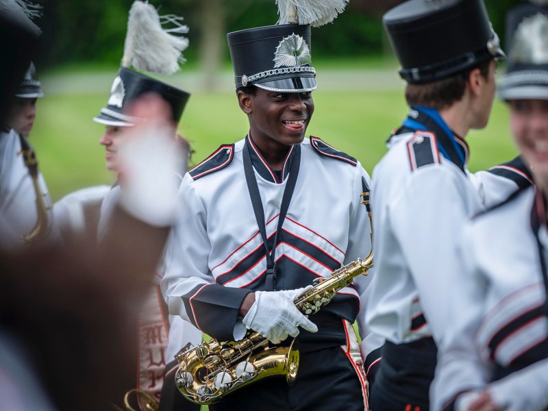 A marching band member in a black and white uniform holds a saxophone and smiles. Others in similar attire are visible in the background. The scene is outdoors with grass and trees.