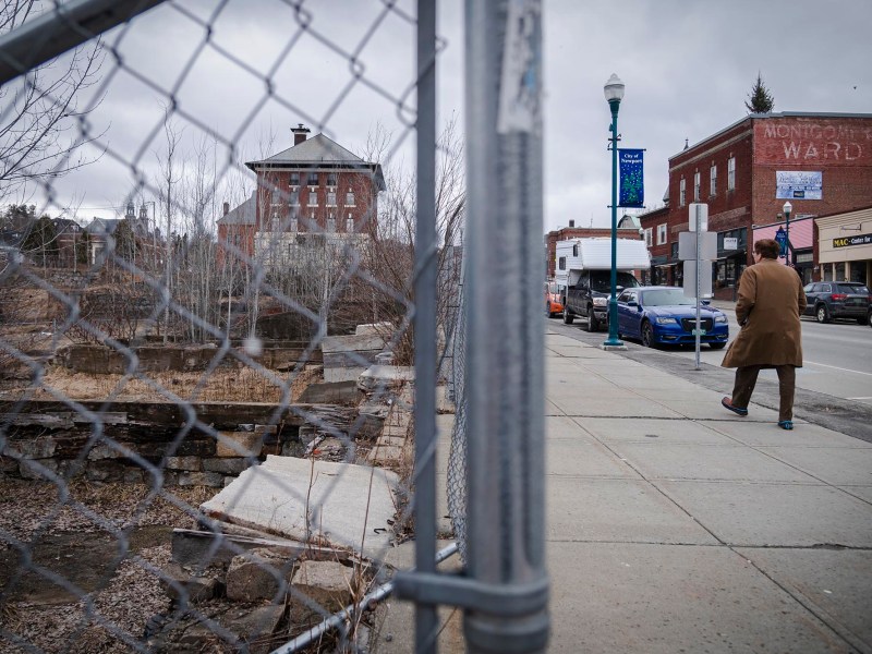 A person walks by a chain-link fence on a city sidewalk, with historical buildings in the background.