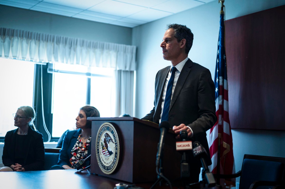A man in a suit speaks at a podium with a microphone, flanked by two seated women, in a conference room with an american flag.
