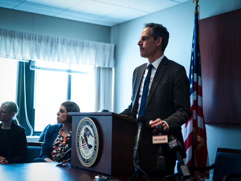 A man in a suit speaks at a podium with a microphone, flanked by two seated women, in a conference room with an american flag.
