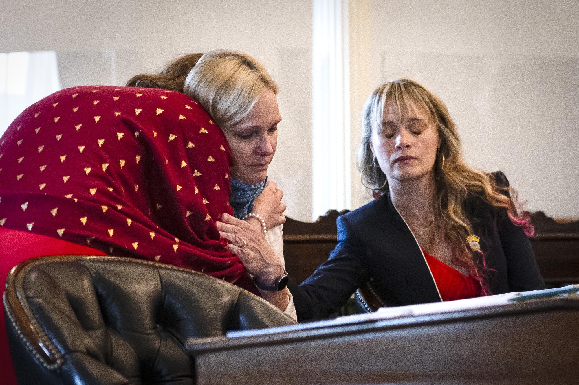Three women in an indoor setting, one wearing a red patterned headscarf leaning in, another blonde woman in the center, and a third woman with wavy blonde hair and a black blazer on the right.