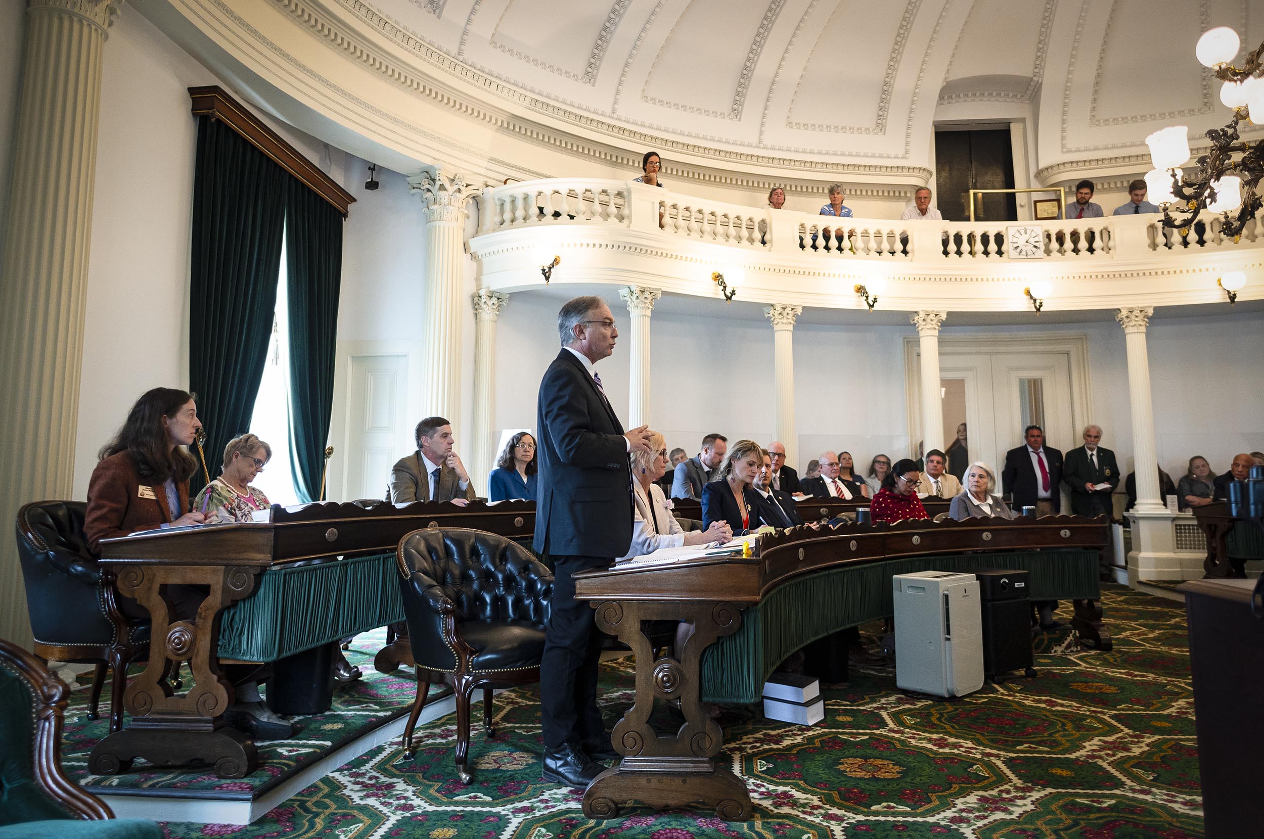 People are seated and standing in a formal, circular room with ornate decor, likely during a government or legislative session.