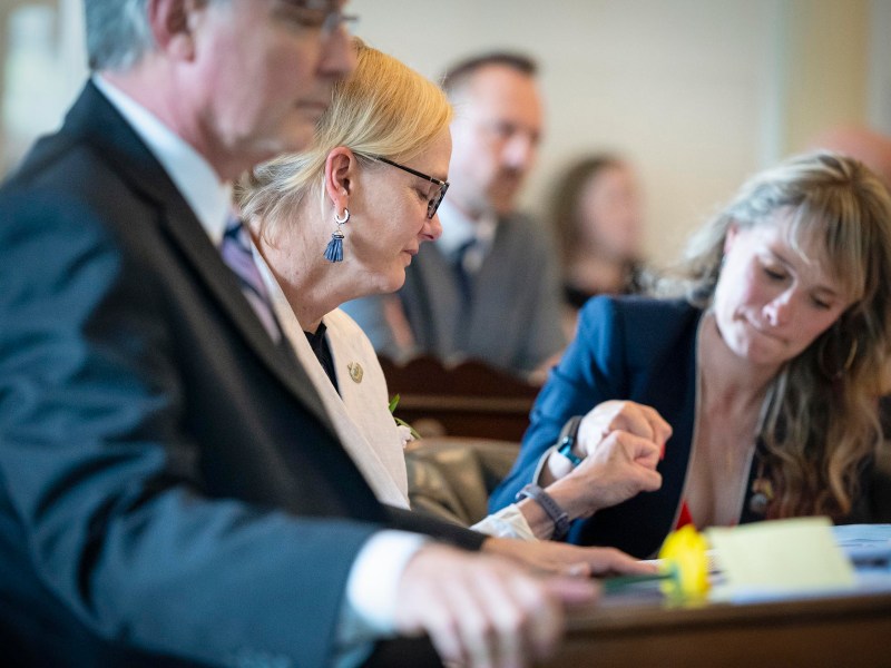 A woman in a light blazer and glasses interacts with another woman in a dark blazer while seated at a table, with other people sitting and standing in the background.