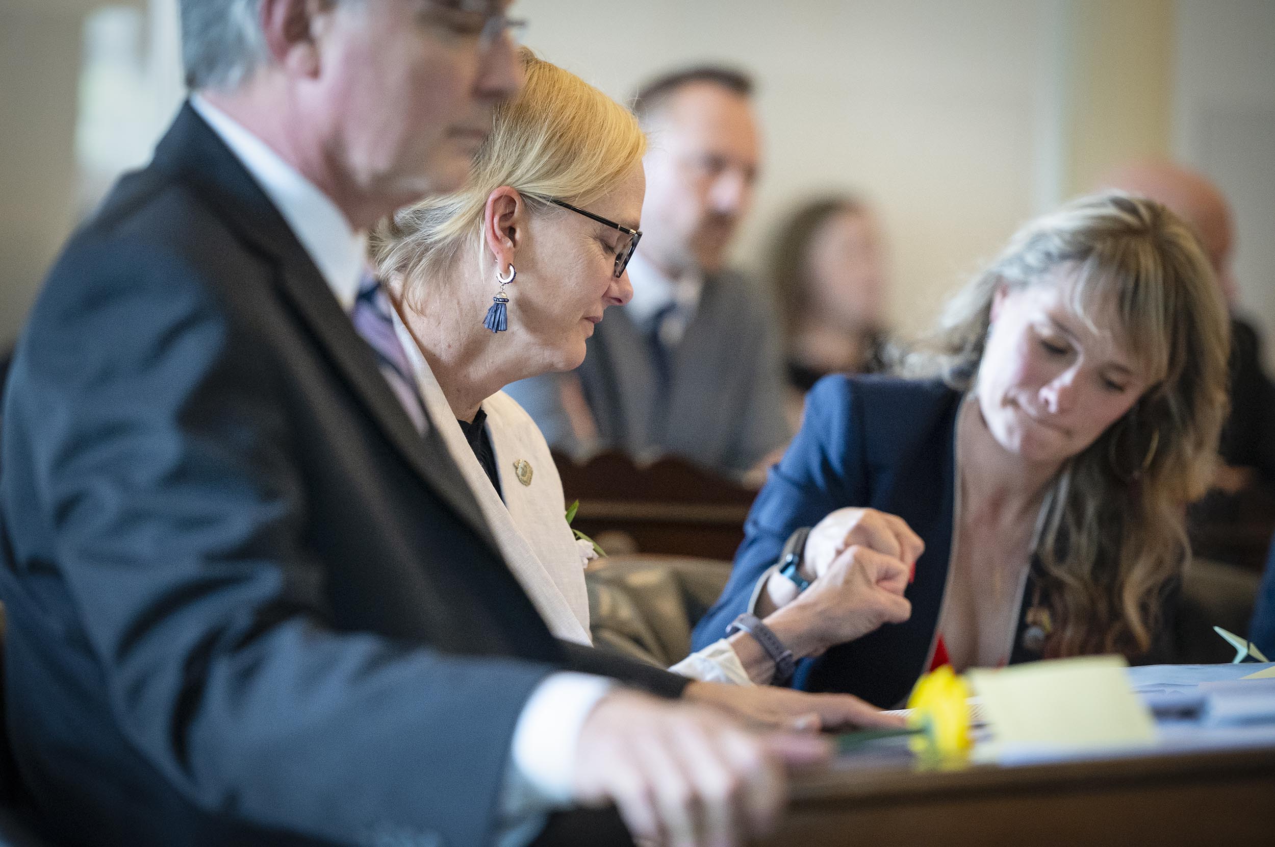 A woman in a light blazer and glasses interacts with another woman in a dark blazer while seated at a table, with other people sitting and standing in the background.