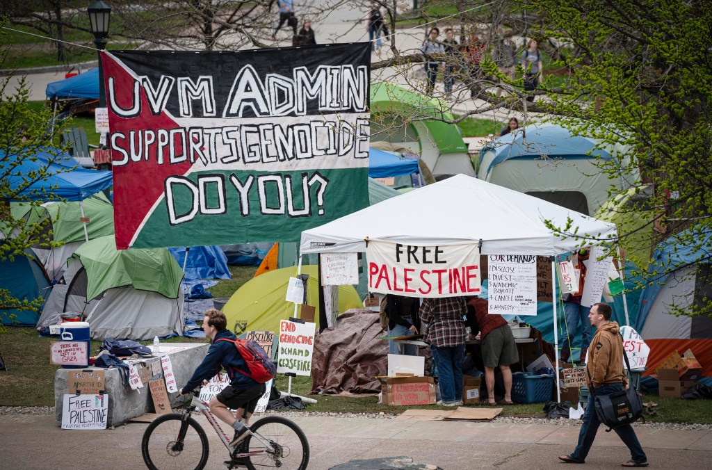 A protest camp with banners stating "uvm admin supports genocide, do you?" and "free palestine," with people walking and biking past.