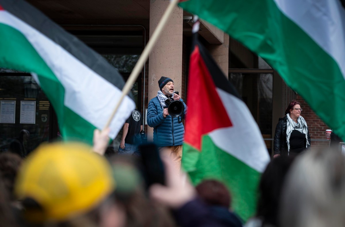 A photographer captures images at a rally with people holding palestinian flags, focusing through a large lens, with a blurred crowd in the foreground.