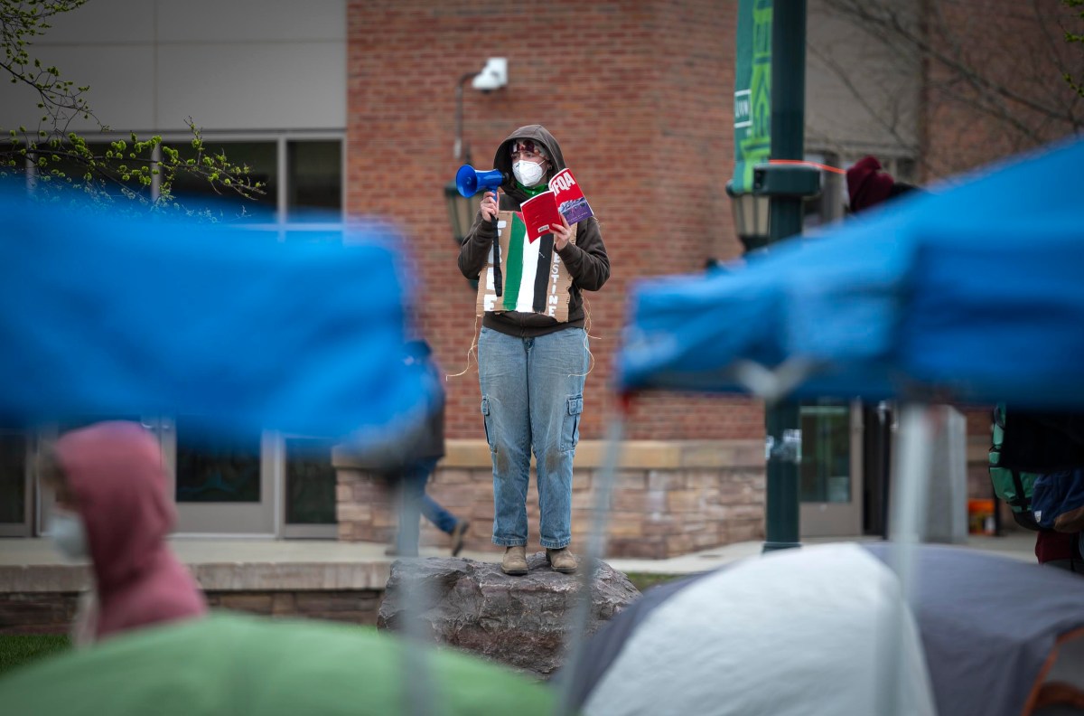 Person standing on a rock, speaking through a megaphone at a protest, holding a sign, with tents and listeners in the foreground.