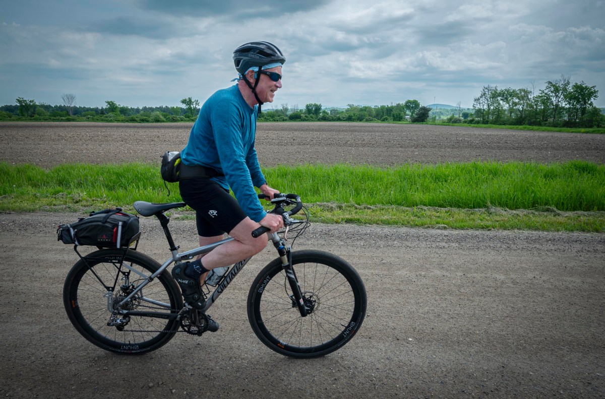 A cyclist wearing a helmet and sunglasses rides a loaded touring bike along a dirt road with greenery and fields in the background.
