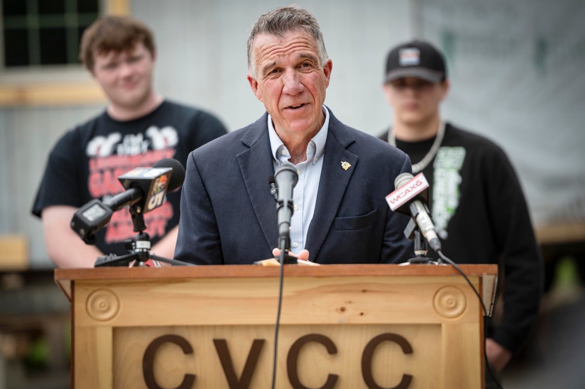 A man in a suit speaks at a podium labeled "CVCC" with two people standing behind him. There are microphones from different media outlets on the podium.
