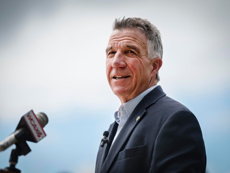 A person in a dark suit speaks at a podium with a microphone, standing against a cloudy sky background.