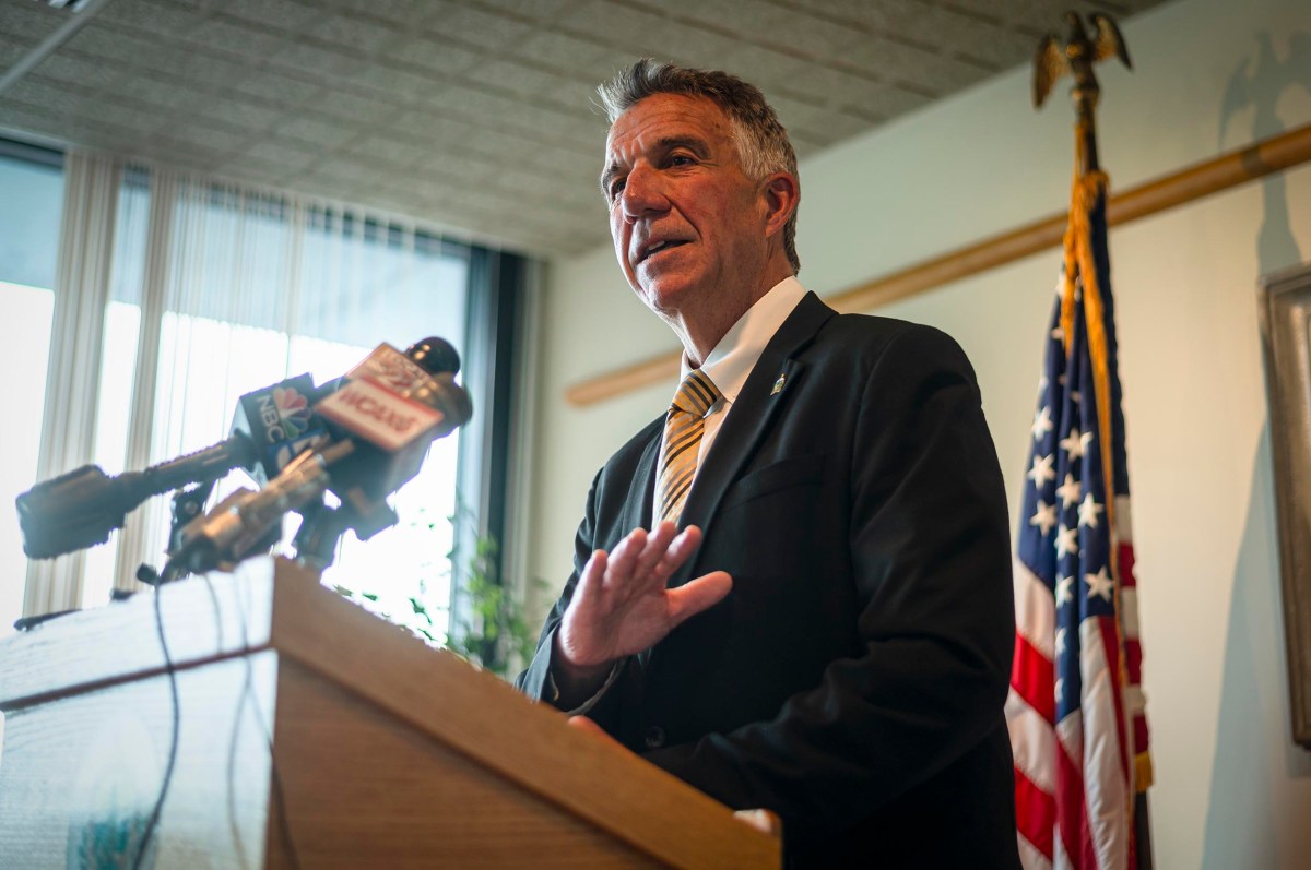 A man in a suit speaks at a podium with microphones in front of the American flag and a window with blinds.