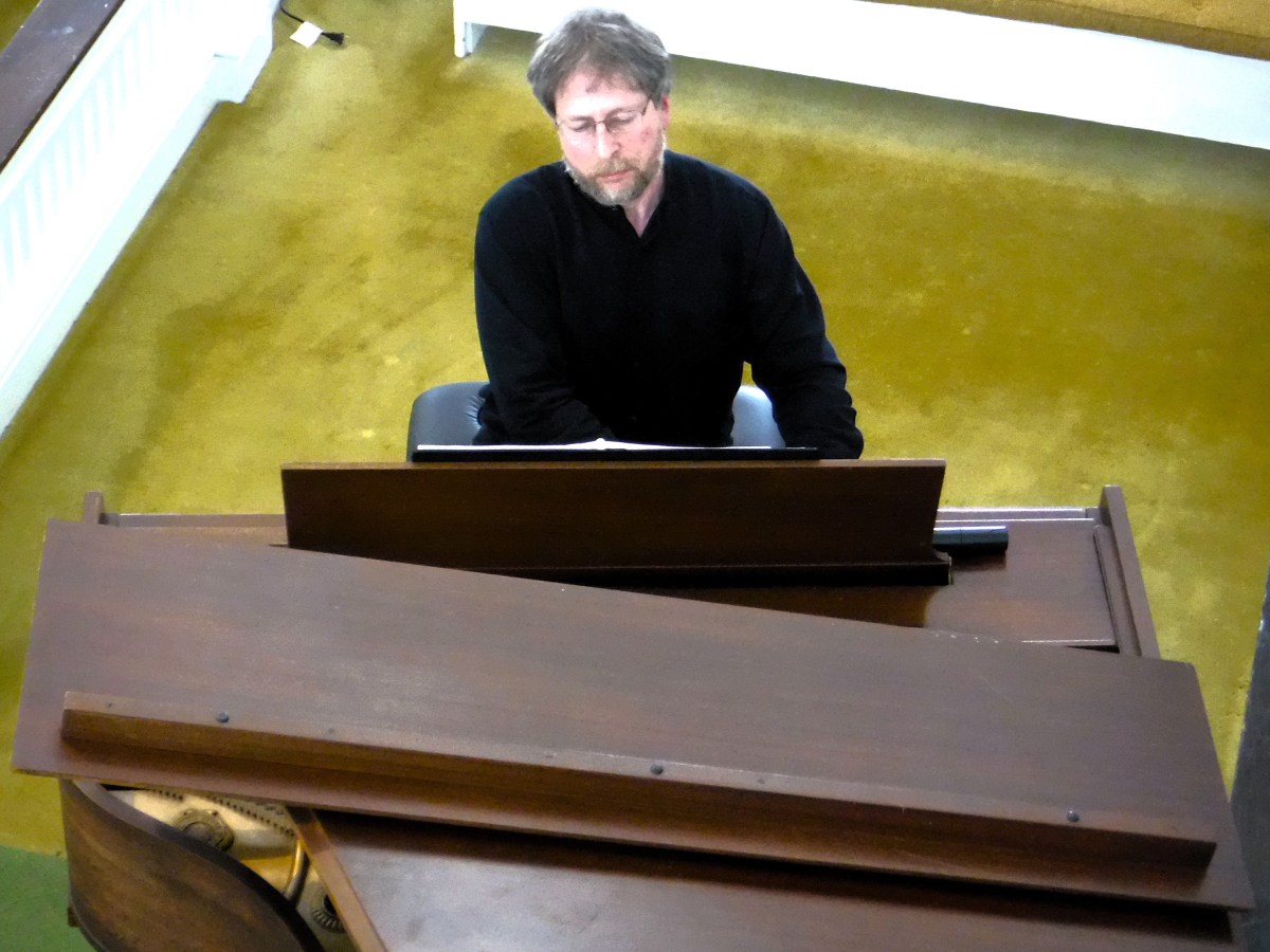 A person with short brown hair and glasses is playing an upright piano, wearing a black shirt, in a room with green carpeting.