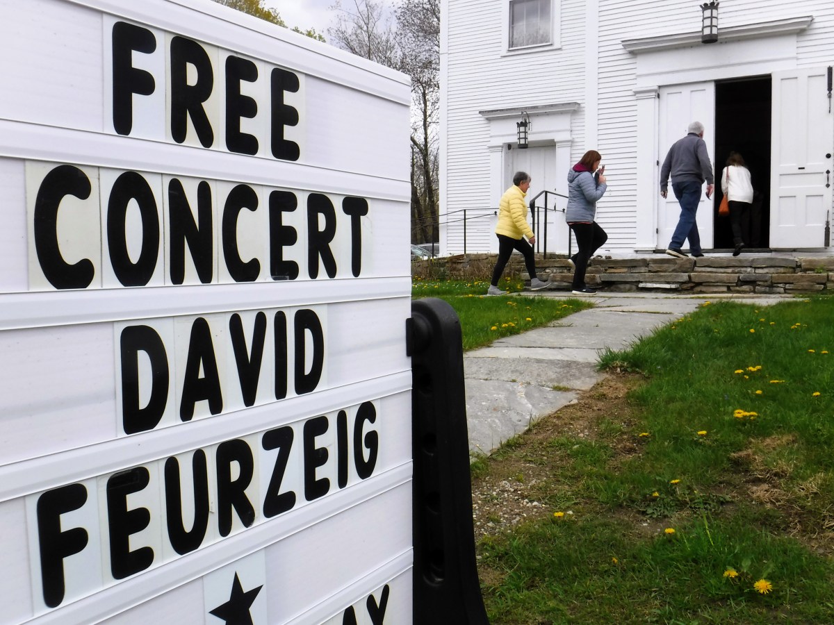 People enter a building for a free concert by David Feurzeig, as displayed on a sign in the foreground.