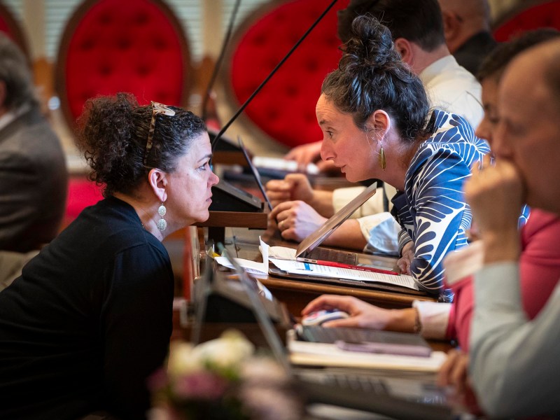 Two women engaging in a discussion at a conference table surrounded by other attendees.