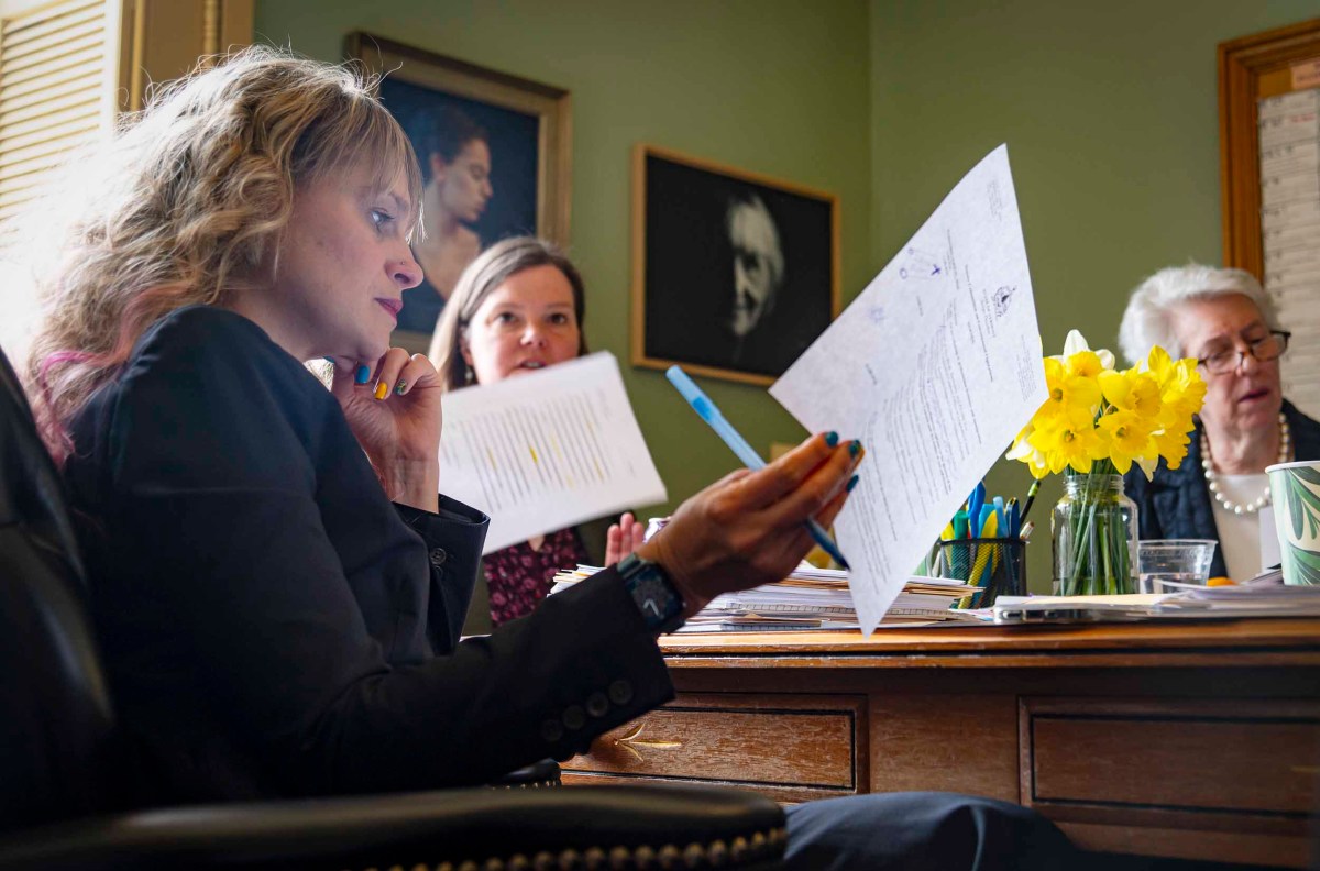 Three women reviewing documents at a desk, one holding a paper while others look on attentively, with yellow flowers in a vase on the table.