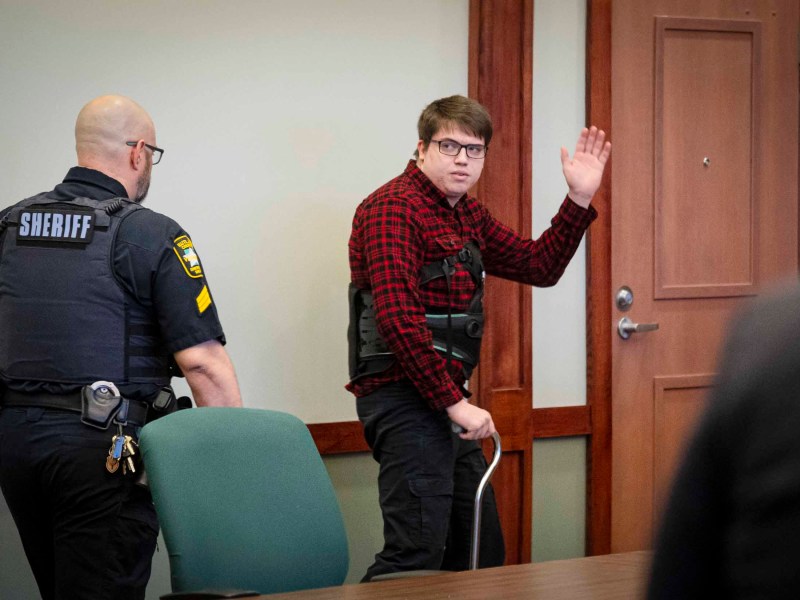 A person in a plaid shirt raising their hand in a courtroom as a sheriff's deputy looks on.