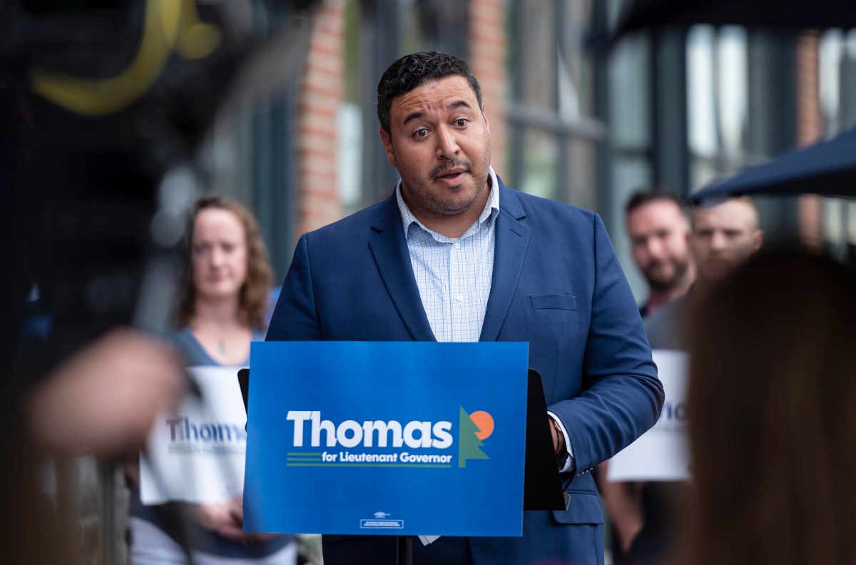 A man in a suit speaks at an outdoor event, standing behind a podium with a sign that reads "Thomas for Lieutenant Governor." People holding similar signs stand in the background.